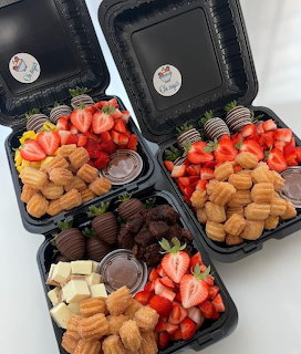 Four black compartment boxes filled with strawberries, churos, chocolate-covered strawberries, and brownies