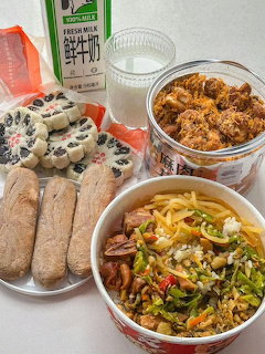 Asian food spread with noodle bowl, fried chicken, rice cakes, sausages, and a glass of milk