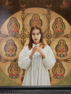 Woman in white robe positioned in front of golden Buddhist mandala with seated Buddha figures