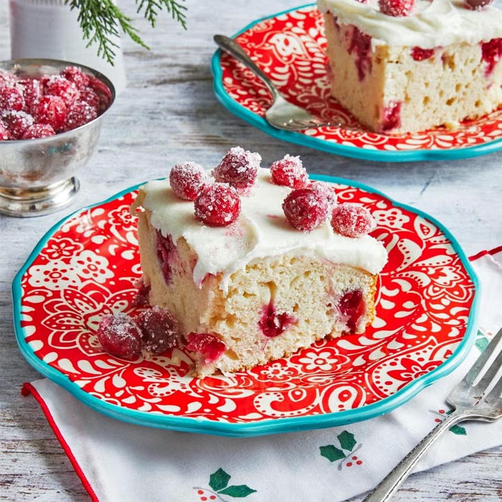 Slice of cranberry cake topped with white icing and sugared cranberries on a red patterned plate