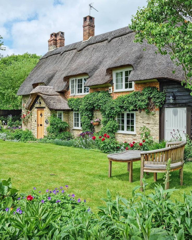 Quaint thatched roof stone cottage with green garden, colorful flowers, and wooden garden furniture