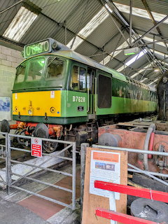 Green and yellow diesel locomotive D7628 parked inside a railway engine shed with maintenance equipment