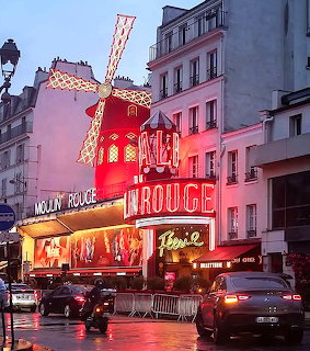 Illuminated Moulin Rouge cabaret on a rainy Paris street at dusk with cars and the red windmill