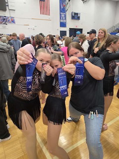 Three girls in costumes and casual wear posing with blue ribbons in a gym