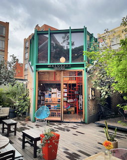 Small glass-front bookstore entrance in a leafy courtyard with benches and potted plants