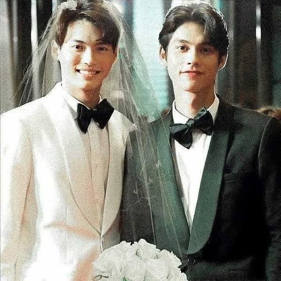 Two men in tuxedos posing indoors with a white bouquet