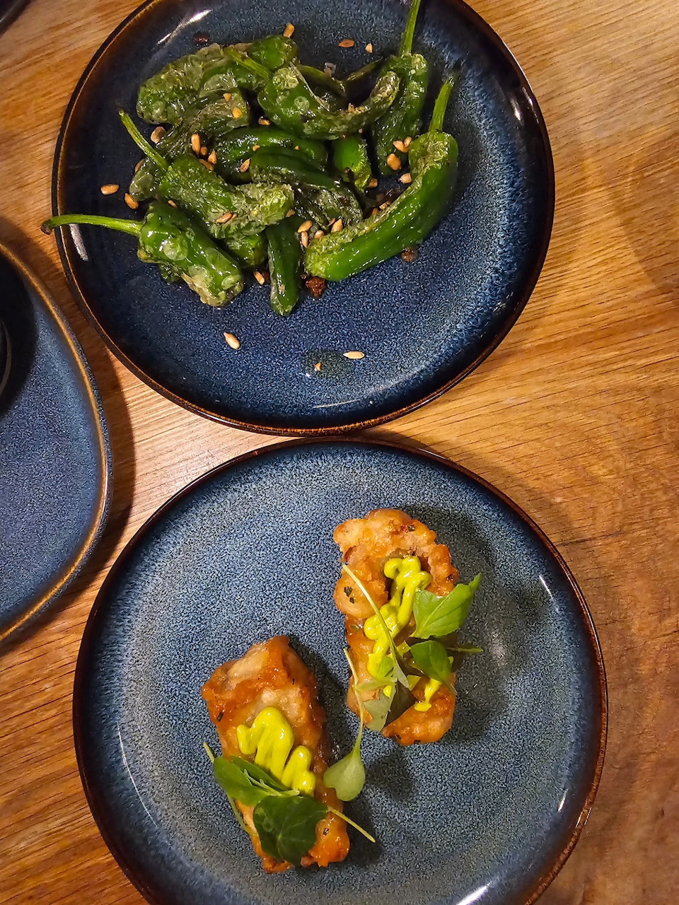 Two dark blue ceramic plates with sautéed green peppers and small garnished appetizer bites on a wooden table