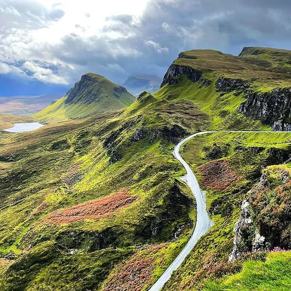 Winding white road cutting through green Scottish Highland mountains under cloudy sky