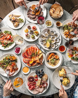 Overhead view of abundant brunch dishes with seafood, meats, fruits, and desserts on white plates with hands reaching