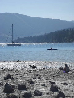 Rocky beach at Sequim Bay with a sailboat, kayaker, and mountain shoreline across calm water.