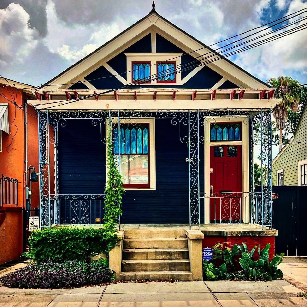 Small navy blue house with decorative white trim, stained glass windows, red door, front porch and garden