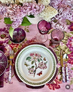 Overhead view of an elegant place setting with botanical plates, purple lilacs, crystal glasses, and floral tablecloth