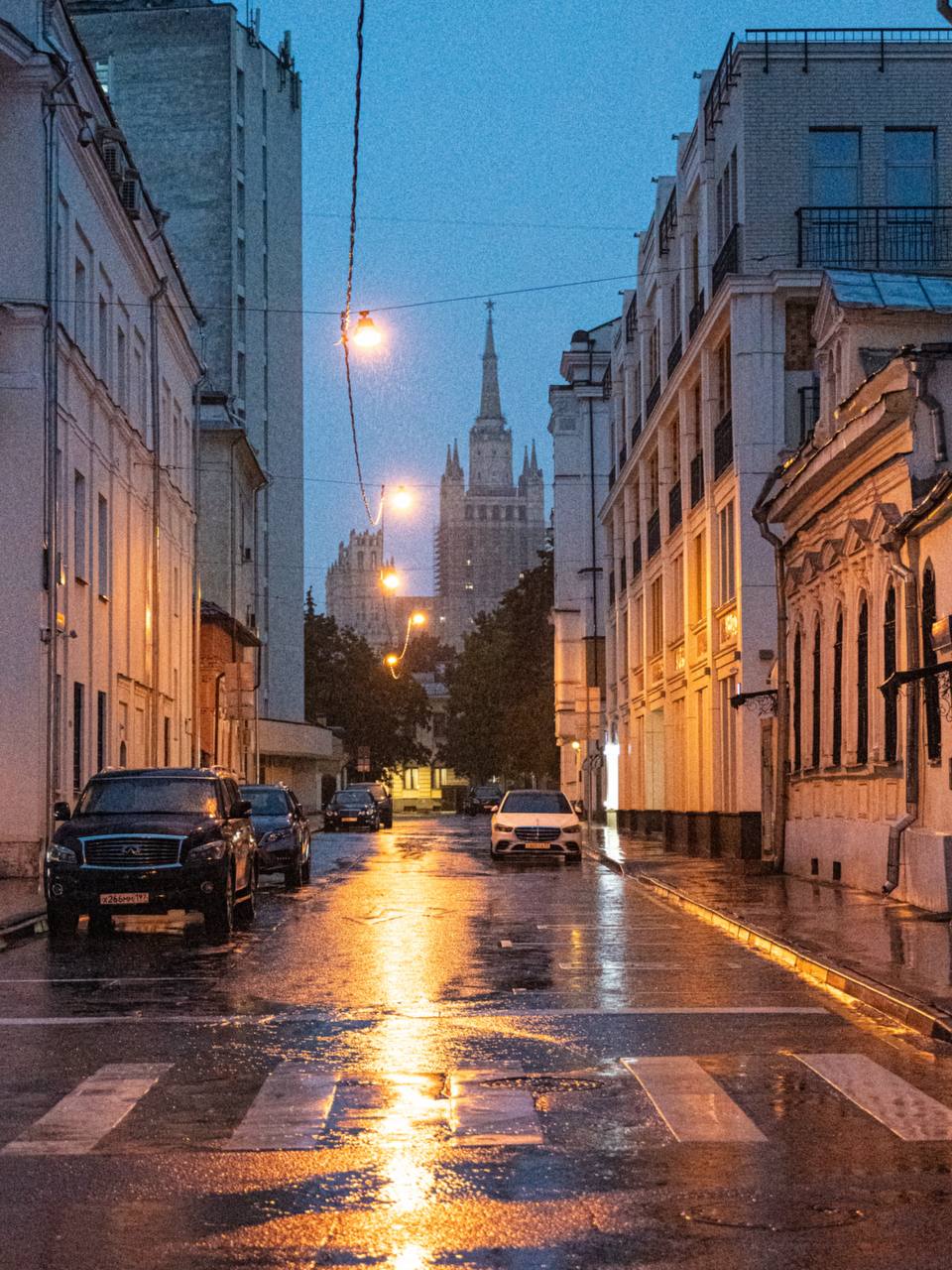 Rainy Moscow street at dusk with parked cars and glowing streetlights leading toward distant spire