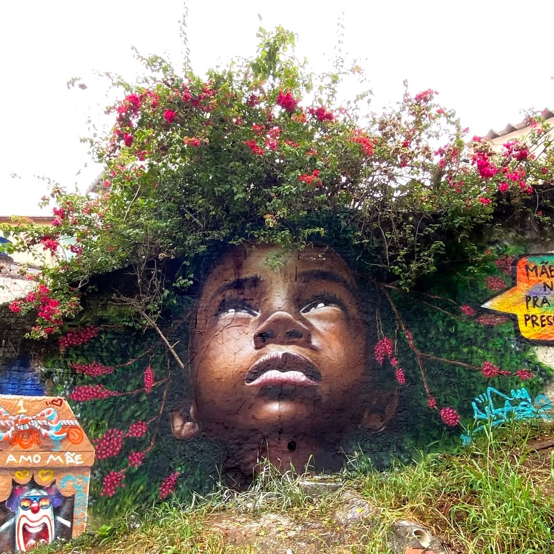 Large mural of face gazing upward with colorful flowers and greenery forming hair, set in urban environment