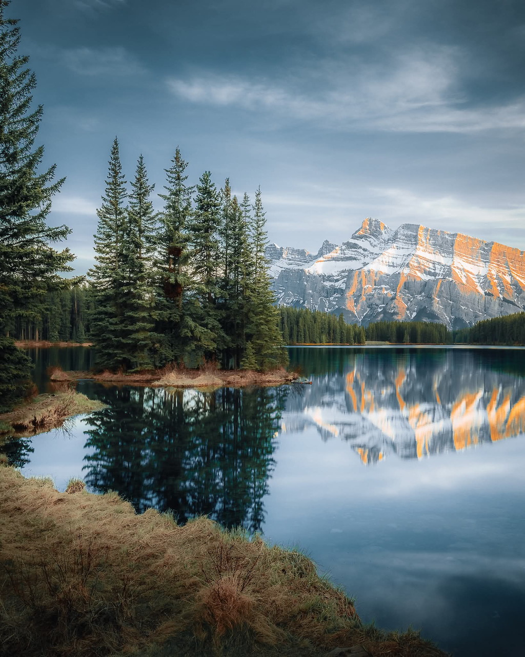 Snow-capped mountain and pine trees reflected in a calm alpine lake under cloudy sky