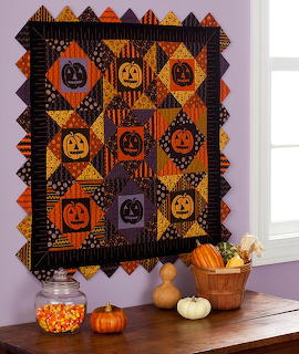 Halloween patchwork quilt with jack-o'-lanterns in orange and black squares displayed on wall with autumn pumpkins