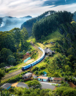 Blue train passing through a green mountainous village with misty peaks in the background
