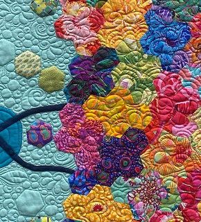 Colorful patchwork quilt with hexagonal floral fabric pieces in bright rainbow hues on light blue quilted background