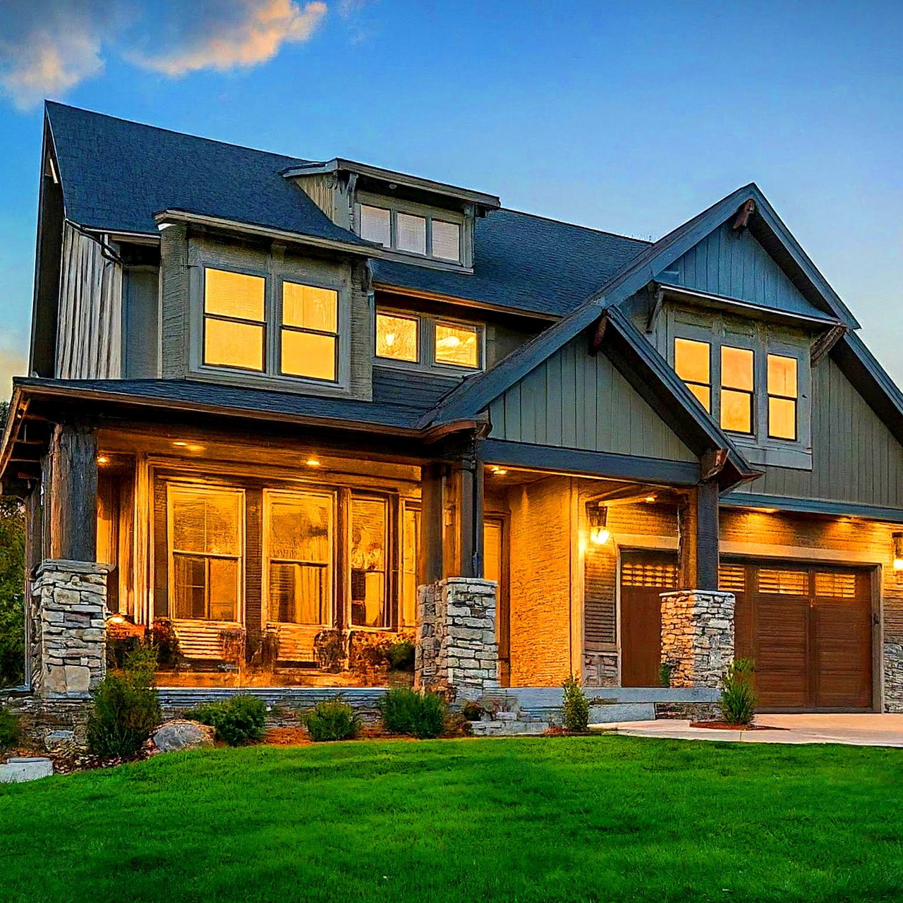 Craftsman style house with warm interior lighting at dusk, stone pillars, dark exterior siding, and green lawn
