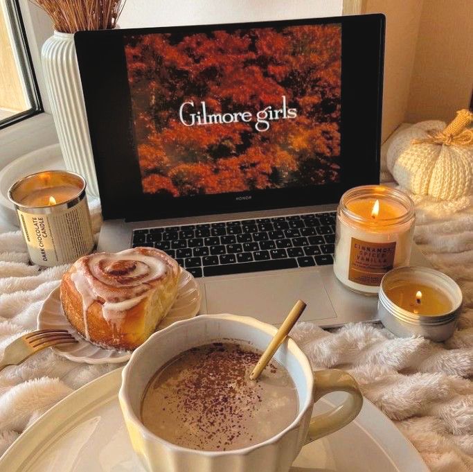 Cozy autumn setup with Gilmore Girls on a laptop, candles, pumpkins, hot cocoa, and a cinnamon roll