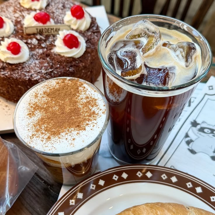 Iced coffee and a frothy coffee drink beside a chocolate cake with whipped cream and cherries