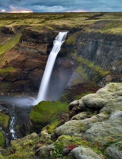 Tall waterfall plunging into a green mossy canyon with dramatic cloudy sky above