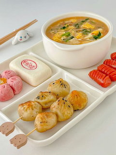 Korean food set with rice cake skewers, steamed bun, pink dessert, tomato slices, and egg soup on white tray