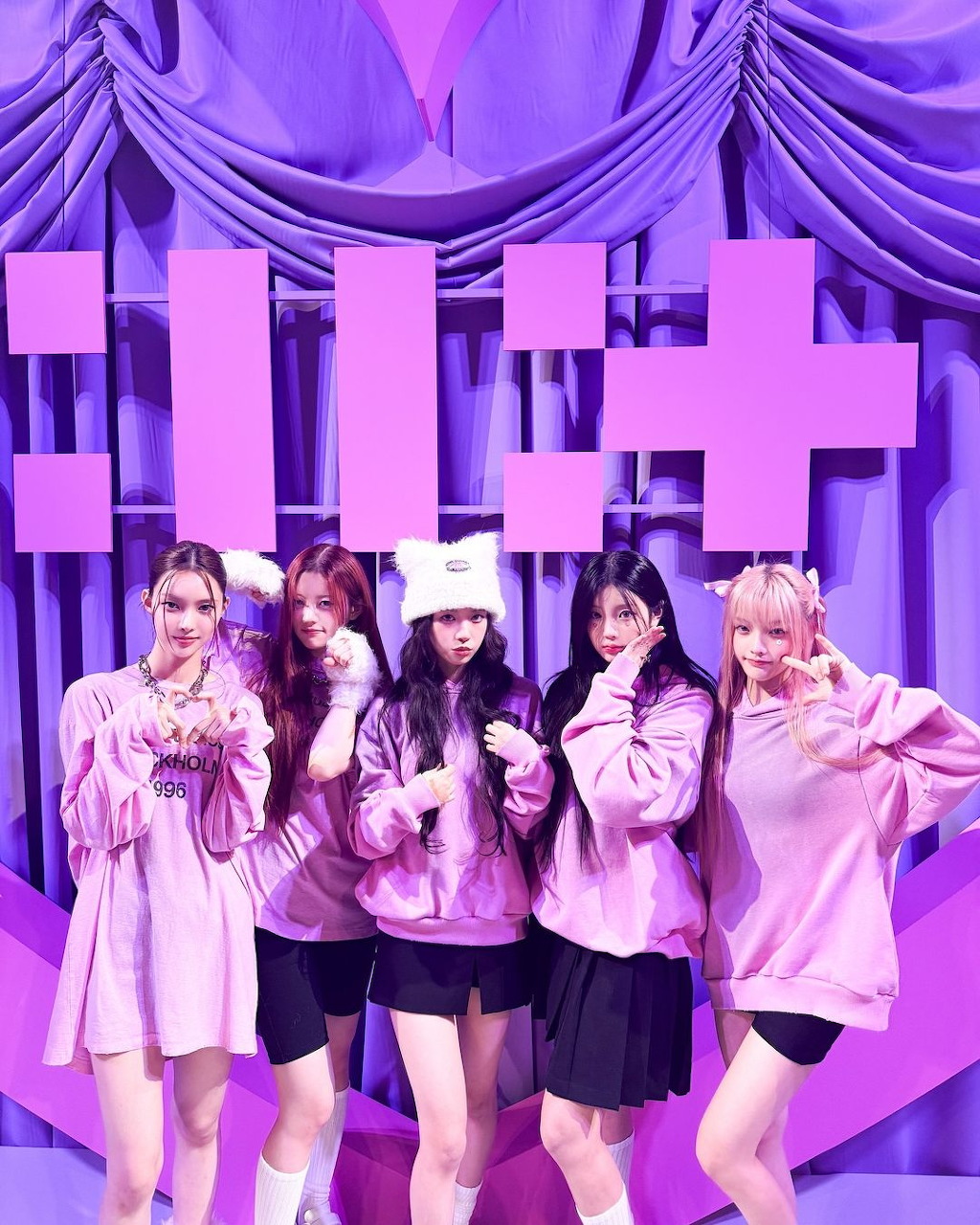 Five young women in pink hoodies posing together in front of a purple backdrop with large letters