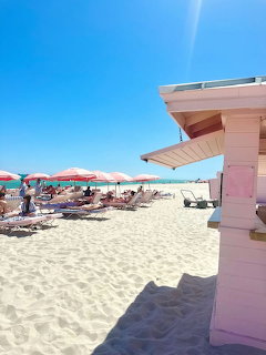Pink beach hut on sandy beach with umbrellas, lounge chairs, and blue sky