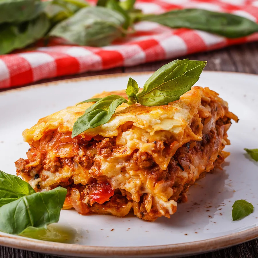Close-up of a slice of lasagna on a white plate with melted cheese and fresh basil garnish.