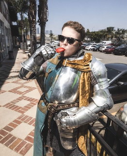 Person in shiny medieval plate armor with gold sash and sunglasses drinking on a sunny sidewalk