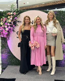 Three women posing on a stage with a pink backdrop and flowers at an indoor event