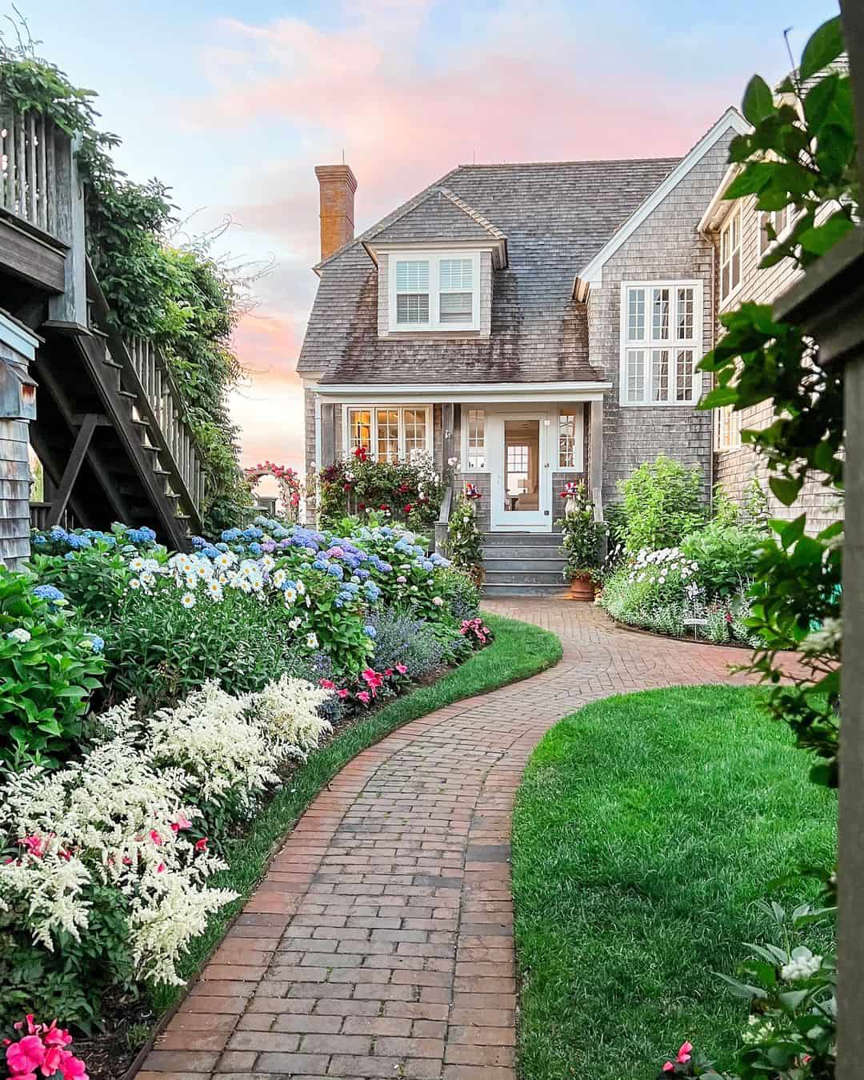 Shingled cottage with brick walkway surrounded by hydrangeas and lush garden at sunset