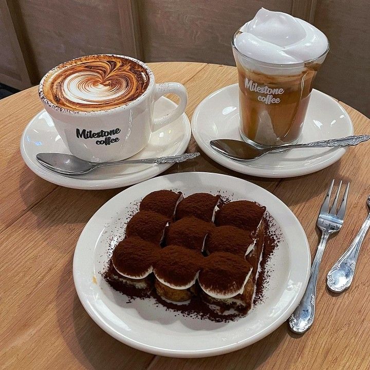 Two coffee cups with foam art and a tiramisu slice on white plates with fork and spoon on wooden table