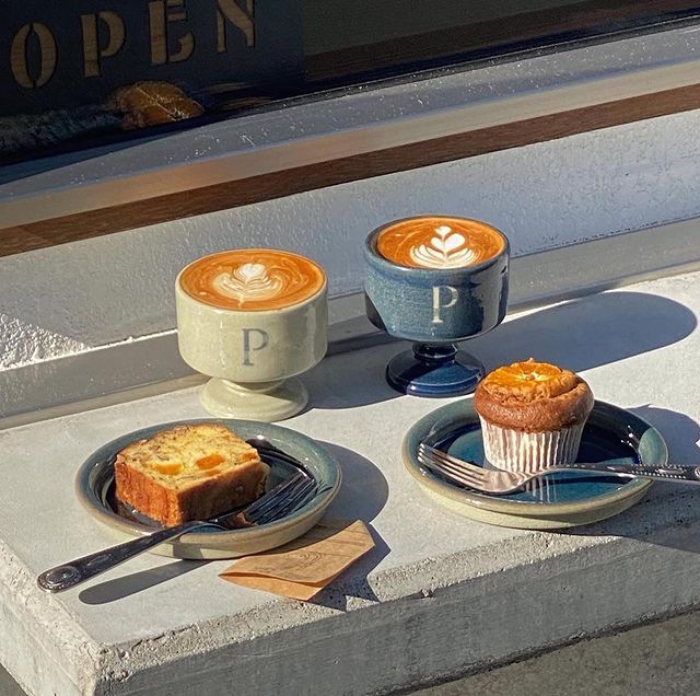 Two lattes with latte art, a muffin, and a toasted pastry on plates in sunlight by a window