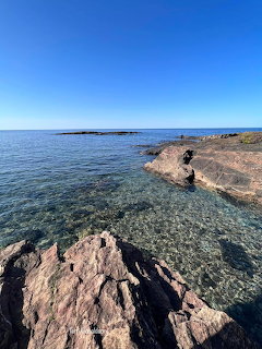 Rocky coastal shoreline with clear shallow water and distant island under blue sky