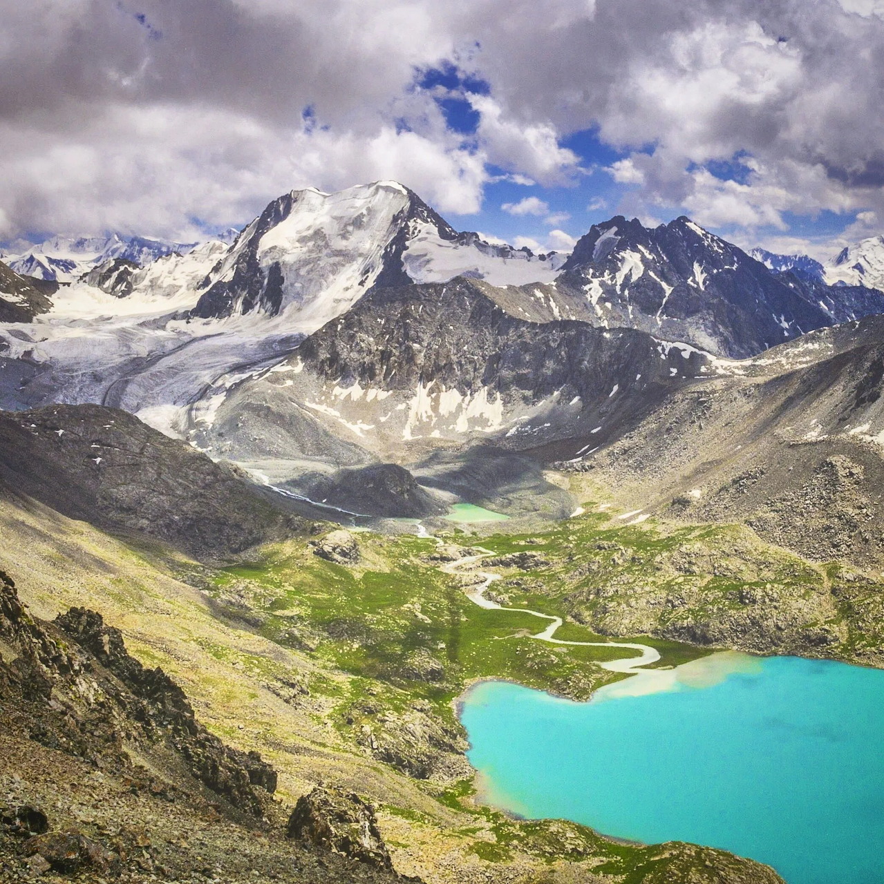 Turquoise alpine lake below snowy mountains and a green valley under cloudy skies