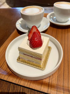 Strawberry shortcake slice on white plate with two cappuccino cups on wooden table