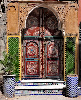 Ornate carved doorway with colorful wooden doors, green tiles, mosaic steps, and two potted plants