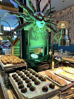 Pastry trays and bread on display inside a decorated restaurant with a large green carnival-style mask