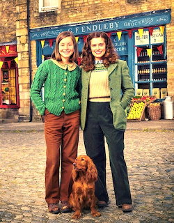 Two women in 1970s clothing pose with an Irish setter on a cobblestone street outside a vintage shop