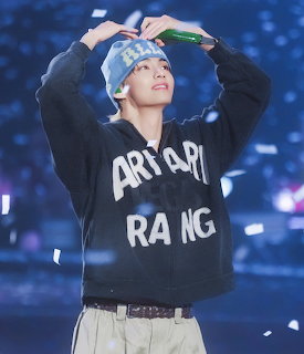Young K-pop idol adjusting blue cap on stage wearing dark hoodie with white text and confetti falling