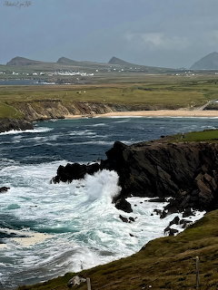 Ocean waves crashing against dark coastal rocks with green fields and distant hills beyond