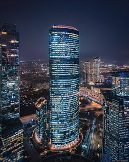 Tall modern glass tower lit up at night with curved architecture surrounded by city buildings and highways