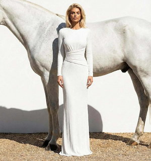 Woman in white dress standing next to white horse in outdoor setting