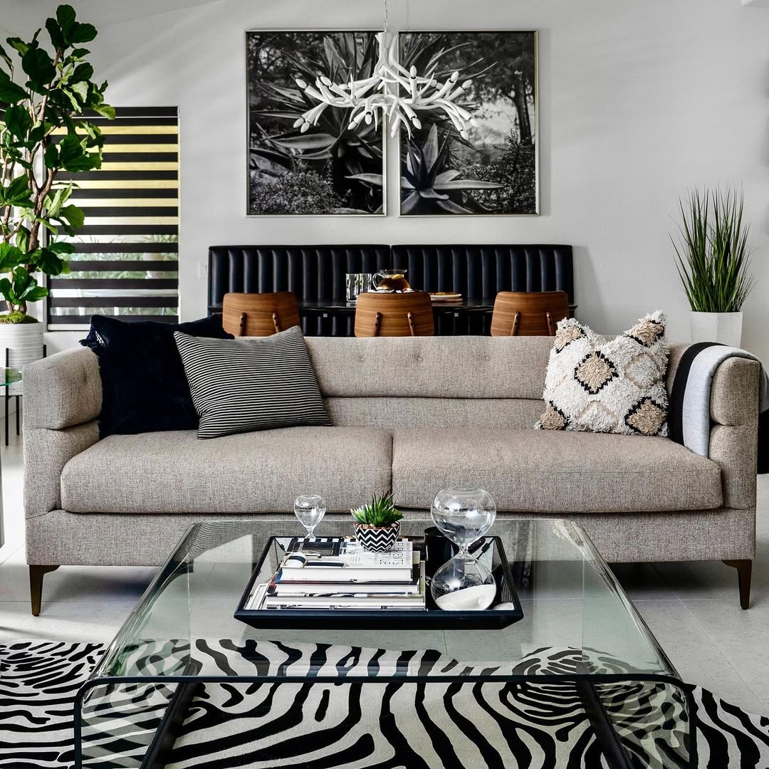 Modern living room with beige sofa, glass coffee table, plants, and black-and-white rug