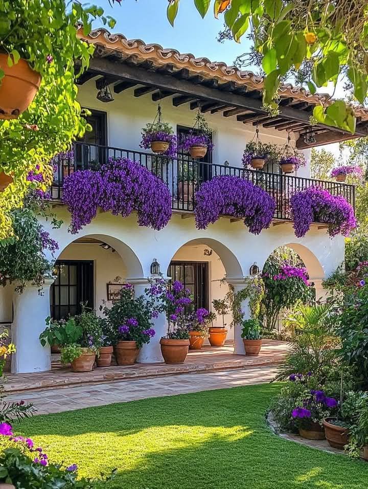White villa with purple flowers cascading from balconies, arched entrances, terracotta pots, and green lawn