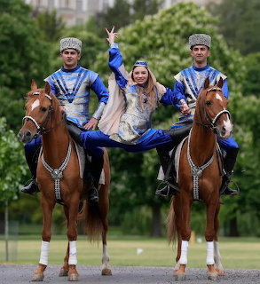 Three riders in blue traditional costumes performing on two brown horses outdoors