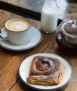 Latte with foam art, cinnamon roll, milk bottle, and teapot on a wooden table