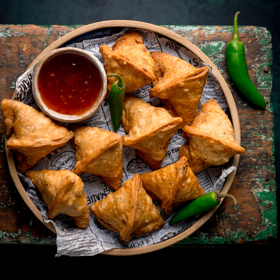 Plate of golden samosas with red dipping sauce and green chilies on a rustic surface